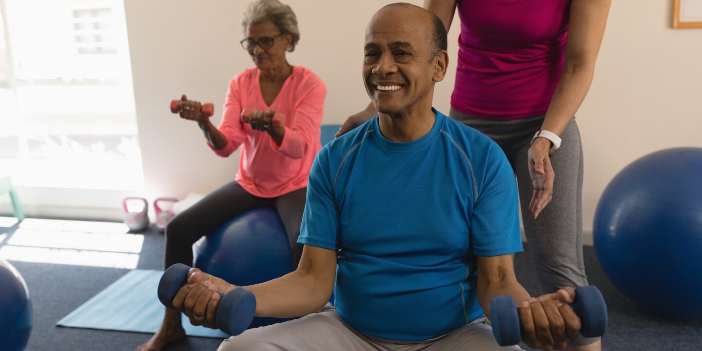 Young female trainer assisting senior man making exercices on exercice ball in fitness studio