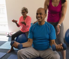 Young female trainer assisting senior man making exercices on exercice ball in fitness studio