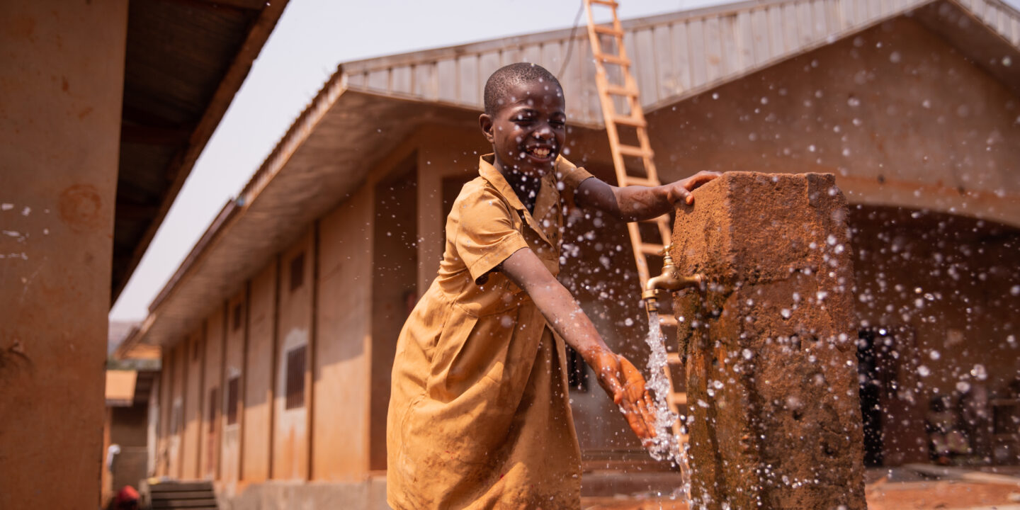 African schoolchild plays splashing water from the tap placed outside in the schoolyard